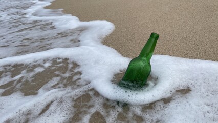 A green glass bottle partially submerged in wet sand on a beach with white ocean foam receding around it.