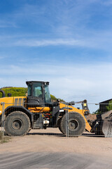Bright yellow wheel loader stands behind metal fence on dusty construction ground. Sunlight highlights large tires, cabin and bucket while green hill and buildings form calm industrial backdrop