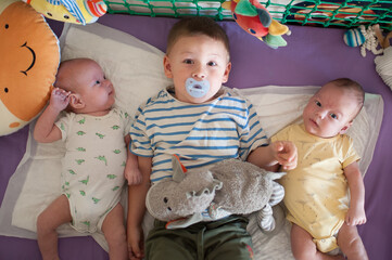 Three brothers are lying on a blanket in a play area. The older brother holds a toy while the younger siblings are next to him. They are surrounded by soft toys and bright colors