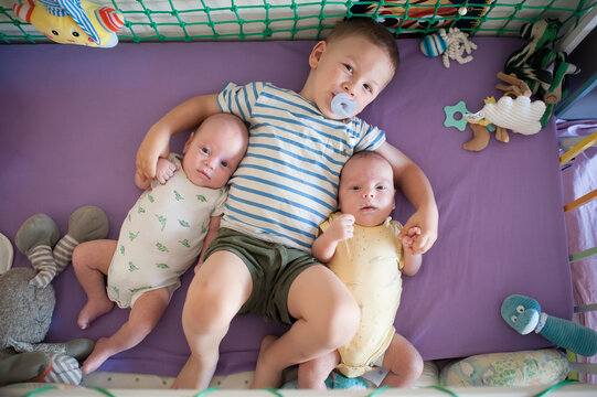 Three brothers rest on a soft blanket in a crib. The older brother lies between the twins and holds a pacifier. Toys surround them as they play and relax - Powered by Adobe