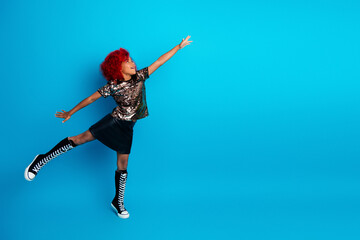 Obraz premium Young girl with red hair wearing a shiny glitter top black skirt and high knee boots jumps joyfully against a bright blue background in a playful fashion moment