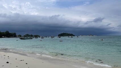 A tropical beach with turquoise water, anchored boats, and distant islands under a cloudy sky.