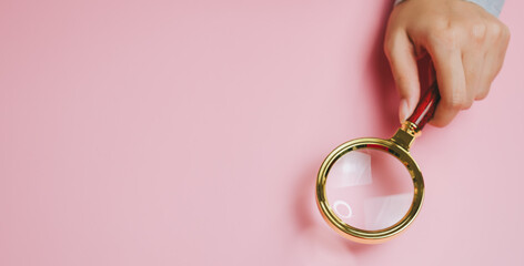 Magnifying glass with golden frame held by hand on pink background, symbolizing focus and detailed...