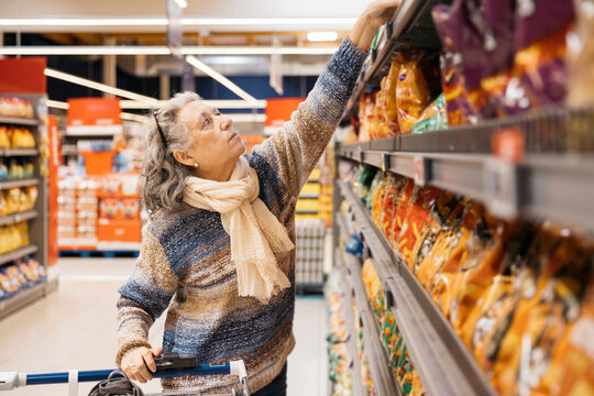 Senior woman reaching for snacks in supermarket aisle