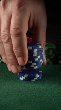 A hand is stacking blue poker chips on a green felt table while other colored chips are nearby. This scene shows a moment from a card game in a busy casino setting at night