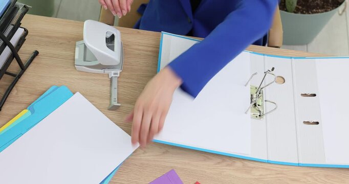 Woman office worker sits at desk and carefully uses hole punch to create neat holes in sheet of paper. Female secretary slides paper into ring binder