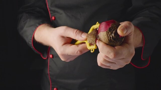 A person holds a beet in one hand and a yellow peeler in the other hand. The background is dark, and the focus is on the preparation of the vegetable in a kitchen.
