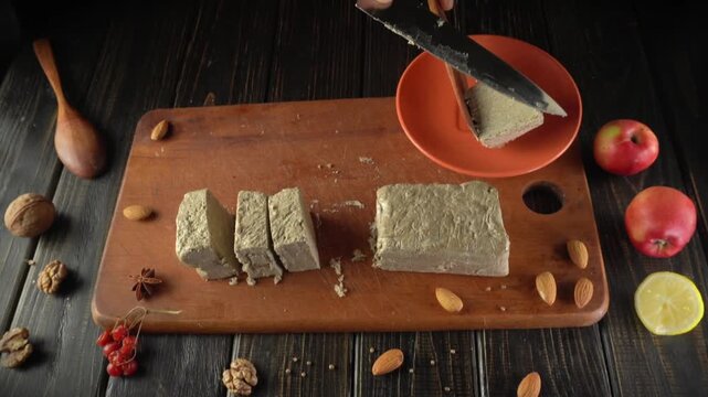 A person cuts a block of sweet treat or halva using a knife on a wooden board. Various nuts and fruits are placed around the board. Cooking is happening in a kitchen setting.
