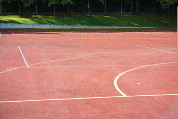Wide view of an empty red outdoor sports court with painted game lines, soft sunlight, and a green tree backdrop creating a calm athletic scene