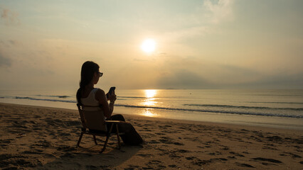Woman sitting on the beach with her phone while watching the sunrise over the ocean.
