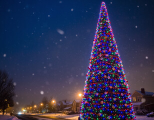 A majestic, brightly lit Christmas tree stands tall in a snowy neighborhood at night, with gentle snowflakes falling. Celebratory and festive. Use for holiday greetings.