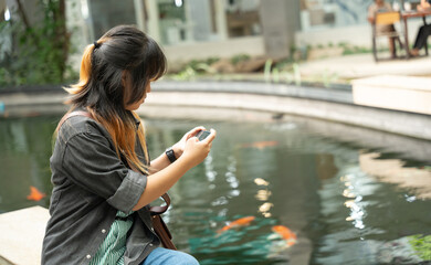 A young girl using her smartphone while sitting beside a koi pond.