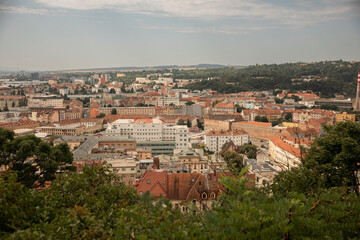 Panoramic city view of Brno from an observation point near Špilberk Castle, showing red rooftops, urban districts and green hills under a soft summer sky in the Czech Republic.
