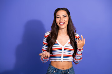 Young woman with long brunette hair wearing striped crop top smiles and points playfully against purple background