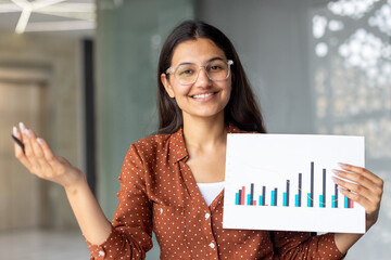 Indian businesswoman standing in an office, smiling confidently and holding up a financial report with a bar chart visualizing data, growth, and development