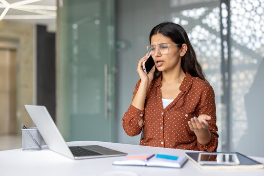 Young indian woman feeling frustrated while having a difficult conversation on her mobile phone, working at an office desk with a laptop and documents