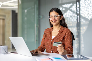 Young smiling indian woman wearing glasses sitting at a desk and using a laptop while holding a...