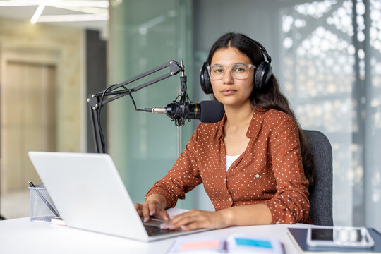 Young biracial woman with headphones and glasses sits at a desk with laptop and professional microphone, focused on recording or editing podcast and online audio content in home studio