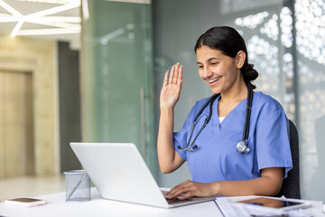 Young smiling female doctor wearing scrubs and a stethoscope making a video call on a laptop, waving her hand during a virtual online medical consultation from her office