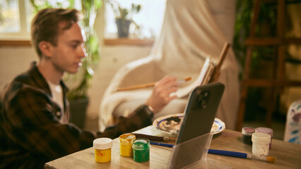 Artist painting with smartphone positioned nearby. Concept of guided creation, following visual cues, flexible learning, and maintaining hands-free workflow in home studio.