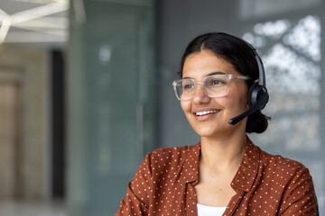 Young woman with headset and clear glasses smiling while working at a call center desk,...