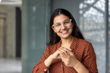 Young indian woman smiling directly at camera, wearing clear frame glasses and a brown polka dot shirt, expressing happiness and confidence in a modern office environment