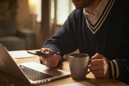 Hand holding smartphone and coffee mug at work desk. Concept of morning routine, multitasking, digital communication, casual workflow, and warm home office moments.