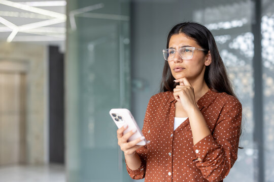 Young indian businesswoman contemplating a challenging problem while looking away thoughtfully, holding a mobile phone in a modern corporate office setting