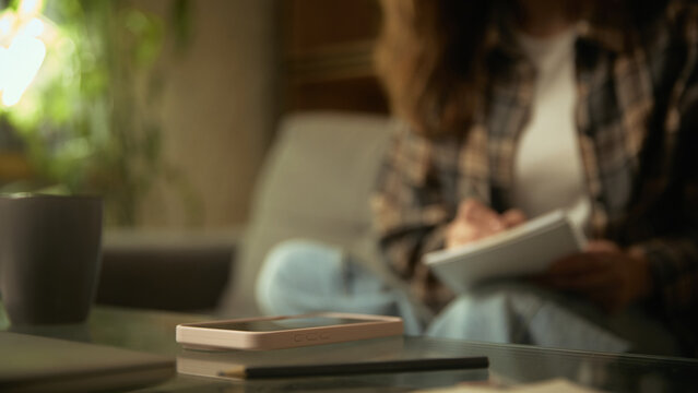 Smartphone lying on table with woman writing in background. Concept of quiet workspace, creative note taking, organized home setup, and digital tools supporting daily tasks.