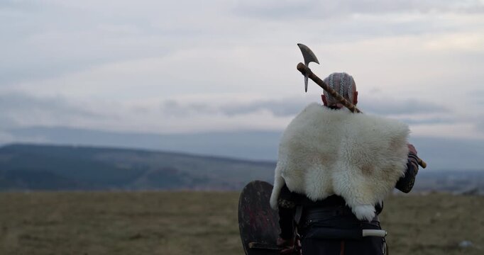 Viking warrior with axe standing on mountain cliff, wearing fur cloak and leather armor, overlooking dramatic landscape. Historical Nordic fighter, strength and adventure concept. Slow motion