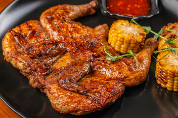 Grilled whole chicken with golden corn on the cob and spicy sauce served on a black plate for barbecue, restaurant menu, and food advertising
