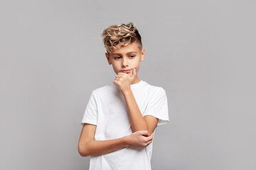 Thoughtful young boy with trendy blond hair touching chin while looking down, wearing white T-shirt and posing in studio, isolated on gray background in modern portrait