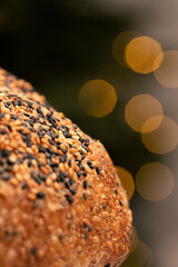 An atmospheric close-up of the curved top of a sourdough loaf, densely coated in a mixture of black and toasted white sesame seeds with a dark background illuminated by warm, orange bokeh circles.