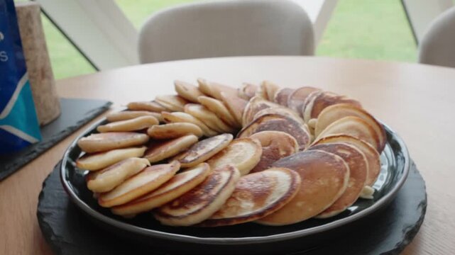 A plate full of small homemade pancakes arranged in a circular pattern on a table. Soft natural light creates a warm and cozy breakfast atmosphere.