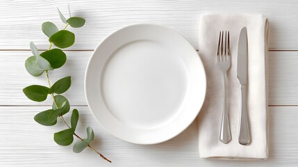 A clean modern place setting featuring a white ceramic plate fork and knife on a beige napkin accented with eucalyptus sprigs on a rustic white wood table background