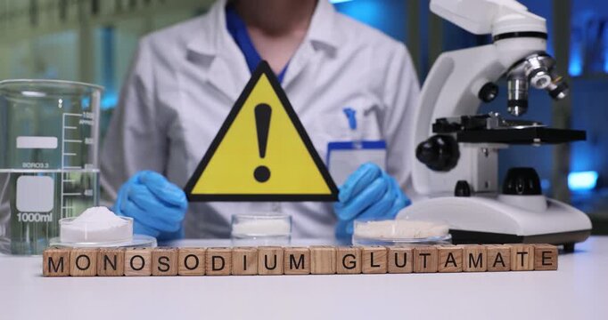 Woman chemist in chemical lab coat holds up warning sign emphasizing importance of safety in laboratory. Mini cubes form phrase Monosodium Glutamate