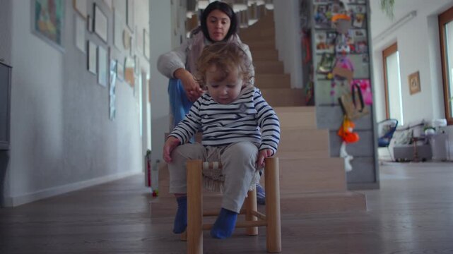 Toddler sitting excitedly on wooden stool while mother watches from stairs creating lively indoor childhood moment shaped by playful movement curiosity safety awareness 