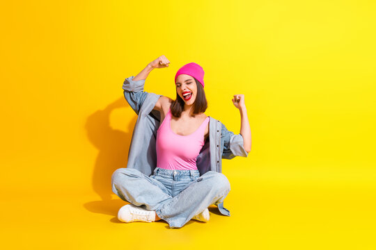 Young woman in pink beanie and denim sits on a bold yellow background flexing arms with a big smile showcasing streetwear style and vibrant fashion energy - Powered by Adobe