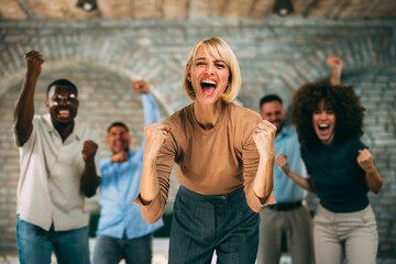 Excited Caucasian businesswoman cheers and raises her fists, celebrating a great achievement with her diverse team in the office