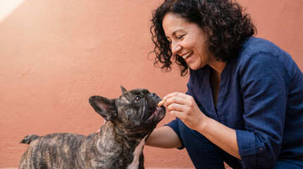 Smiling middle aged woman feeding treat to brindle french bulldog against warm peach wall creating joyful pet bonding moment
