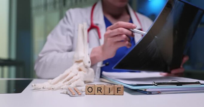 Woman doctor uses X-ray image to inspect bones of foot in details. Wooden cubes arranged on table form abbreviation ORIF near anatomical foot model
