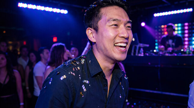Smiling man enjoying vibrant nightclub party with colorful stage lighting, confetti on shirt, and blurred crowd dancing in background