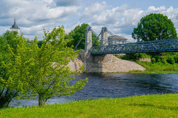 Fototapeta premium Green riverbank with young trees and historic suspension bridge over the Velikaya in Ostrov, Pskov oblast, Russia. stock photo 