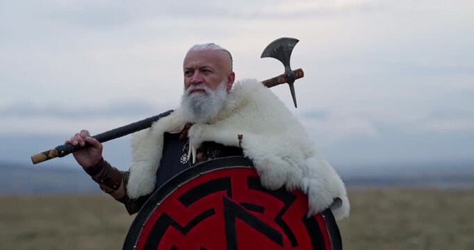 Viking warrior with axe standing on mountain cliff, wearing fur cloak and leather armor, overlooking dramatic landscape. Historical Nordic fighter, strength and adventure concept. Slow motion