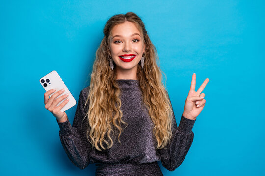 Smiling stylish woman holding a phone and flashing a peace sign against a blue background ready for a festive social media post