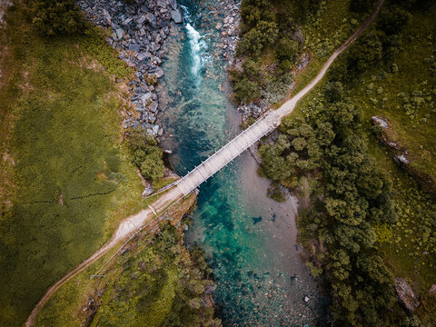 Aerial view of a simple wooden bridge spanning a clear, turquoise river amid rocky banks and lush green vegetation, Rondane, Innlandet, Norway.
