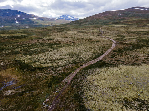 Aerial view of a winding path cutting through the varied textures of Rondane's landscape, contrasting with the distant, snow-capped mountain peaks, Dovre, Innlandet, Norway.