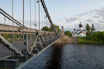 The ancient suspension bridge over the Velikaya River and the Church of St. Nicholas the Wonderworker on a June evening. Ostrov, Pskov oblast, Russia