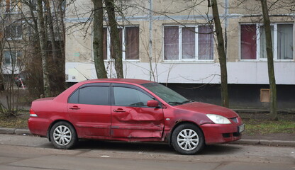 A dented red car is parked near a residential building, Podvoyskogo Street, Saint Petersburg,...