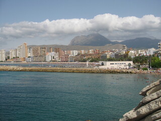 Wide, elevated coastal panorama of Benidorm beachfront, crowded high-rise hotels, urban skyline, and distant rocky mountains under a partly cloudy sky